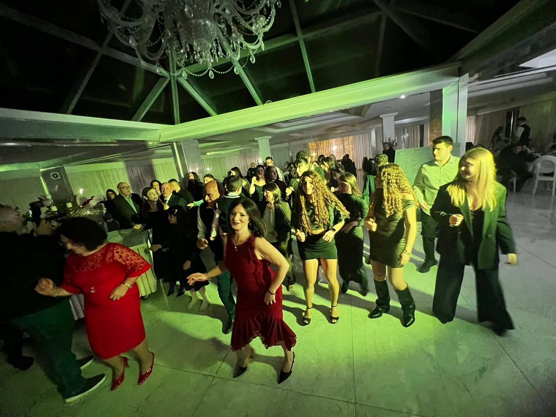 Persone che ballano in una sala da ballo illuminata da un lampadario. La luce verde proietta ombre.