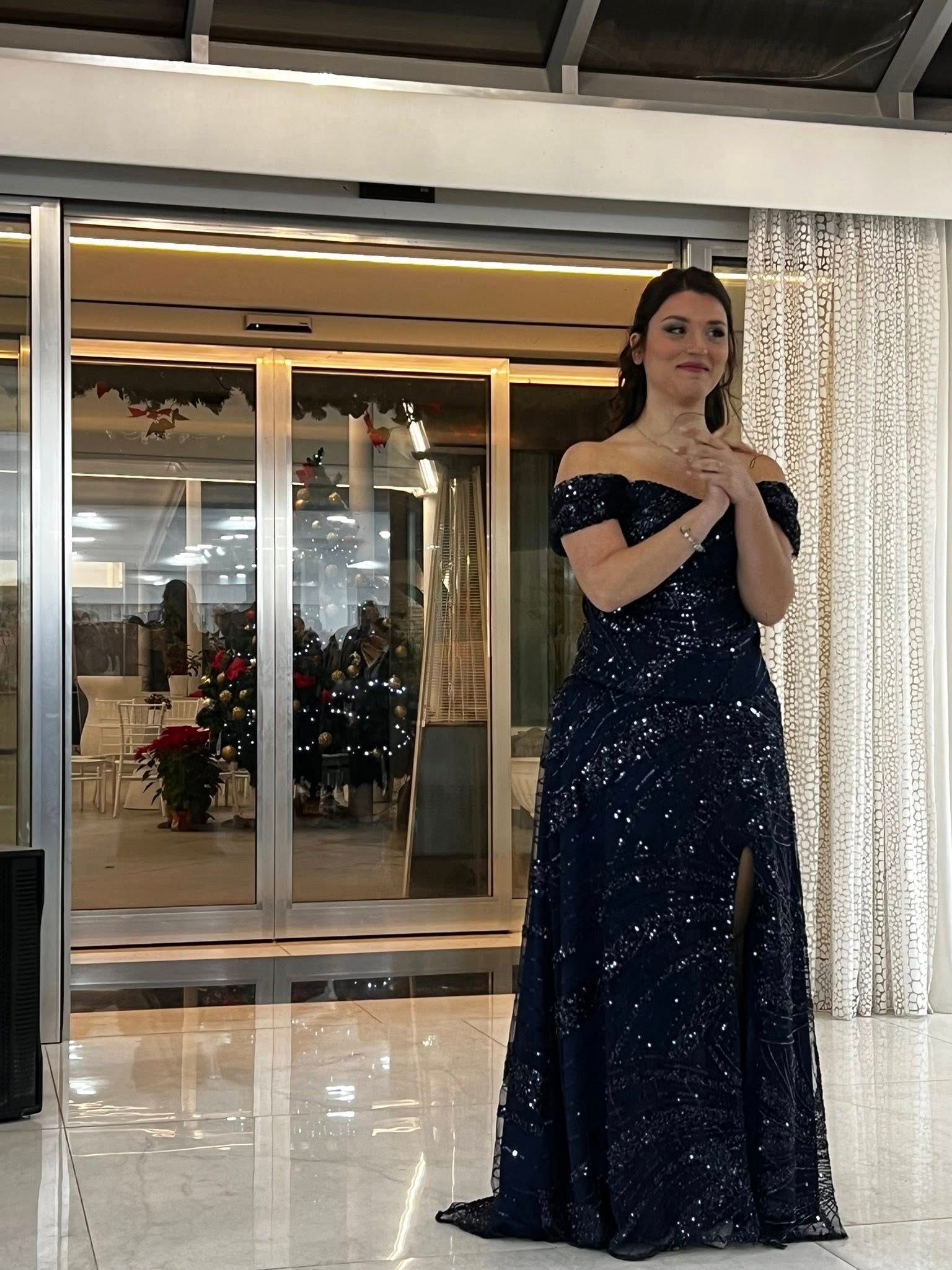 Una donna con un abito blu navy decorato con paillettes è in piedi con le mani giunte davanti a porte a vetri decorate con decorazioni natalizie.