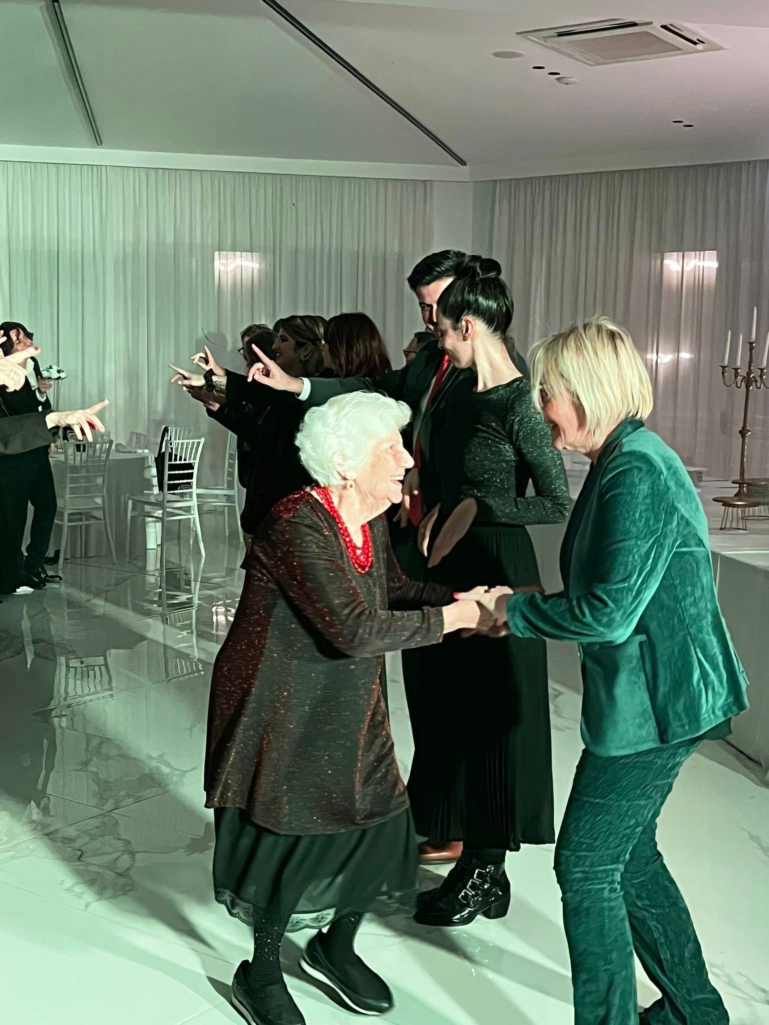 Persone che ballano in una stanza bianca e luminosa, tra cui una donna anziana e un'altra donna in abito verde.