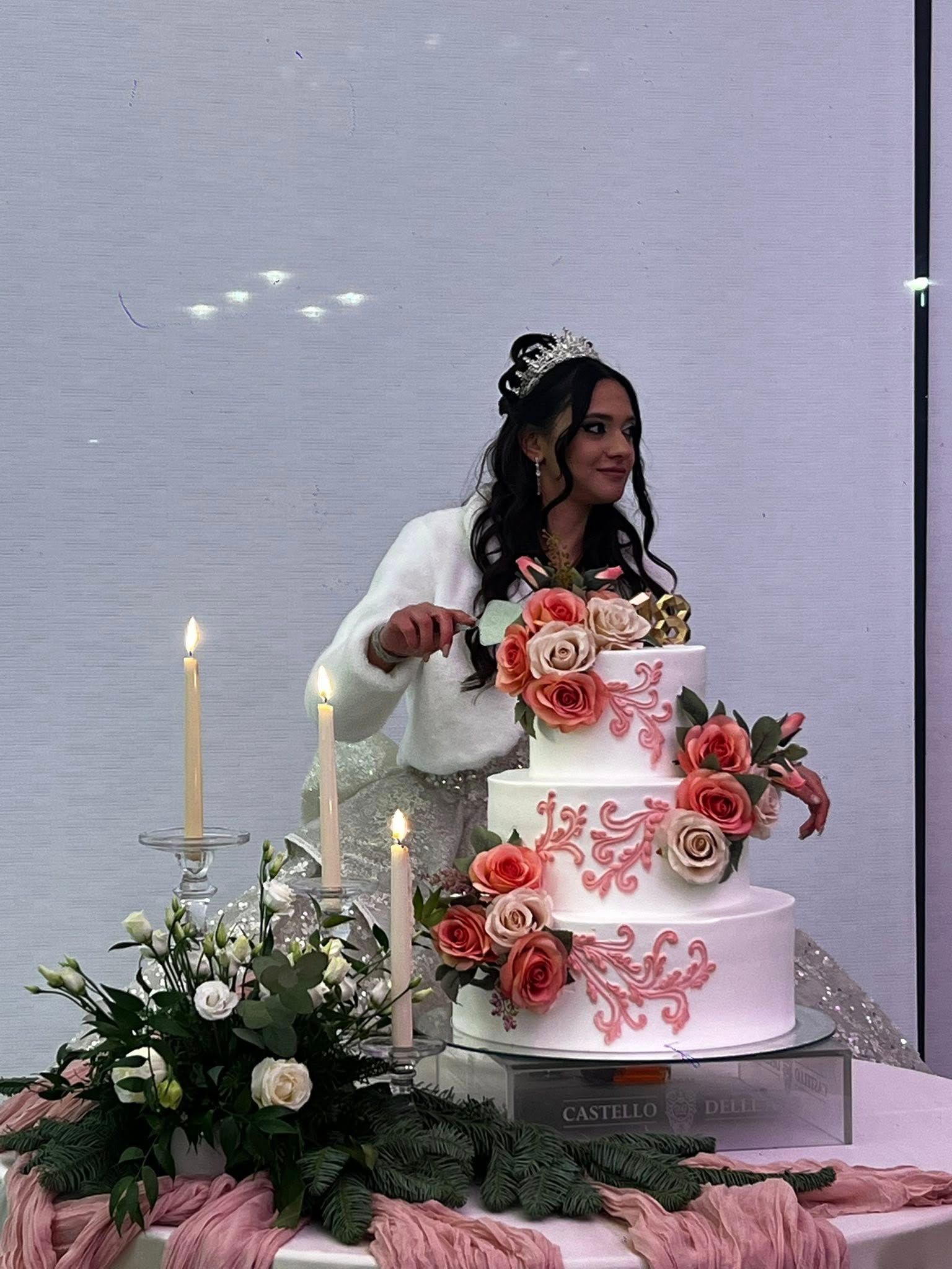 Donna che taglia una torta a più piani decorata con fiori rosa e motivi a spirale. Candele e composizioni floreali sul tavolo.