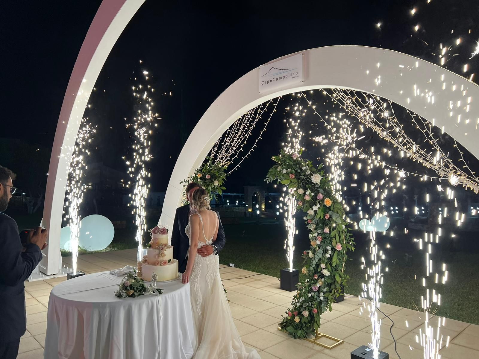 Una coppia taglia la torta nuziale sotto un arco illuminato da bengala di notte.