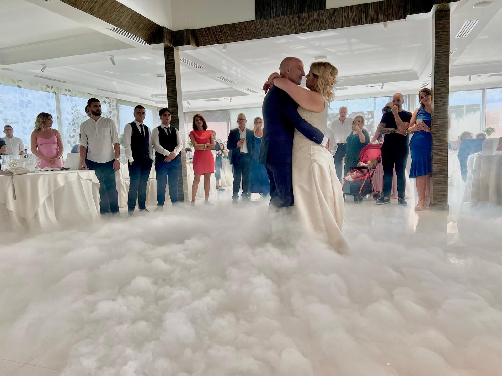 Coppia che balla in una nuvola di nebbia, ricevimento di nozze. Gli ospiti osservano da una sala da ballo con ampie finestre.