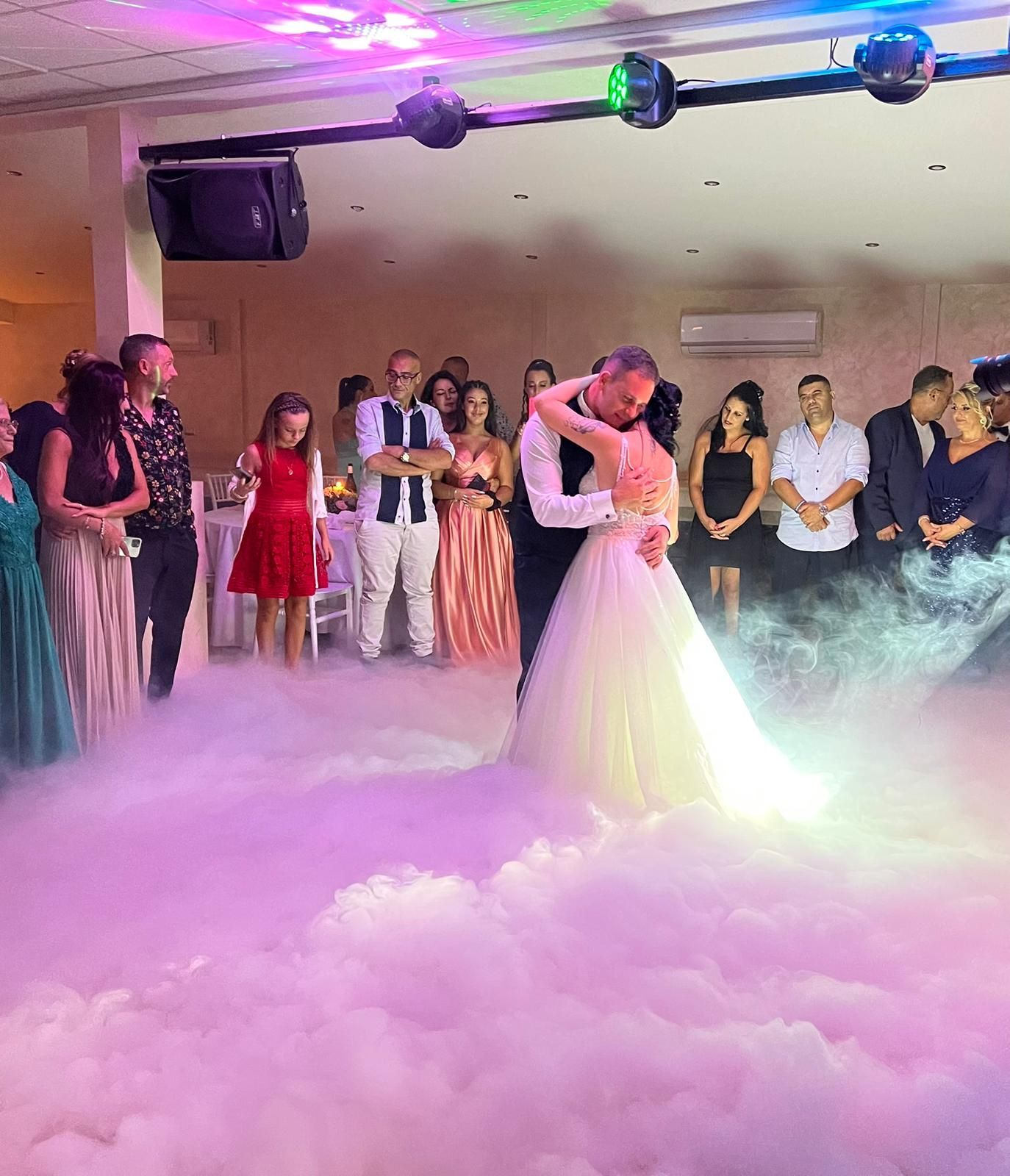 Gli sposi ballano su una nuvola di nebbia durante il ricevimento di nozze. Gli ospiti guardano.