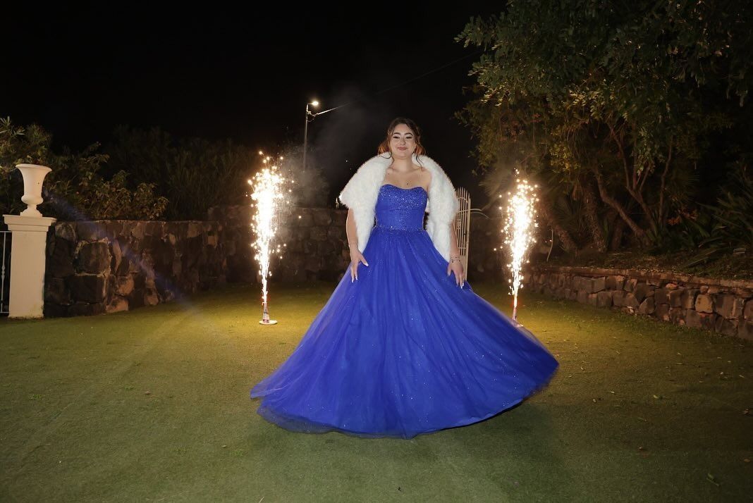 Una donna in abito blu con pelliccia bianca è in piedi sull'erba di notte, mentre i fuochi d'artificio la illuminano.
