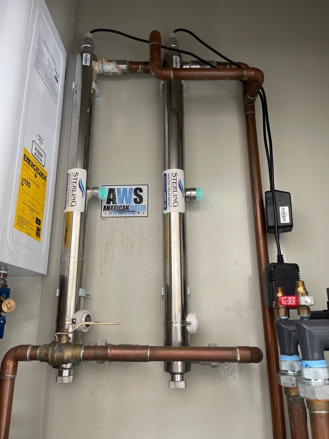 A stainless steel water filter is hanging on a wall next to copper pipes
