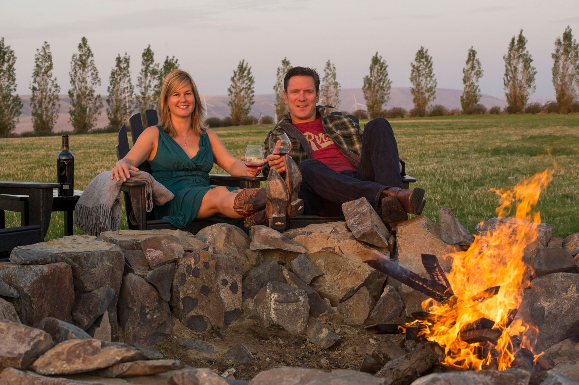 Two people sitting on a rock fire pit beside a campfire in a grassy field at dusk