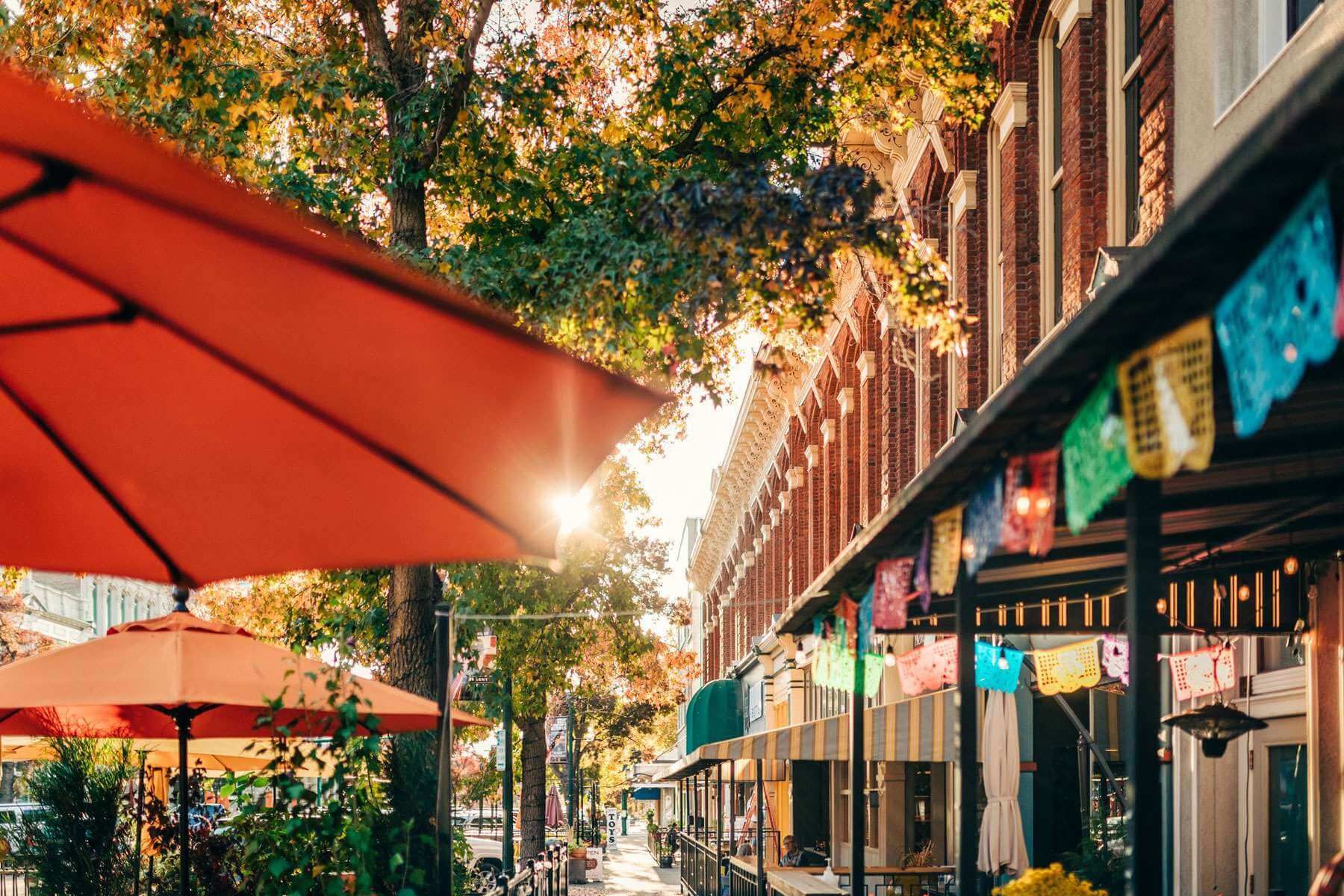 Sunlit street café with red umbrellas, brick storefronts, and autumn trees lining a colorful sidewalk.
