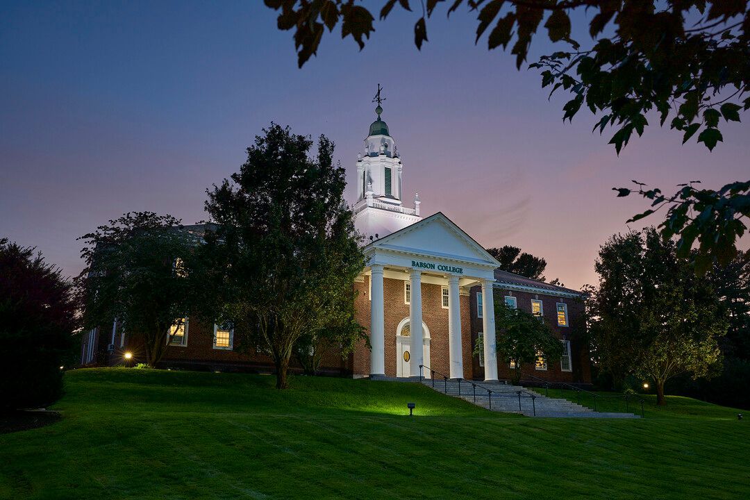 A brick colonial-style building with white columns and a central cupola sits on a grassy hill at twilight.