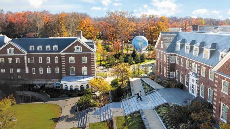 An aerial view of a brick college campus with a large, spherical sculpture, autumn trees, and a person walking on stairs.