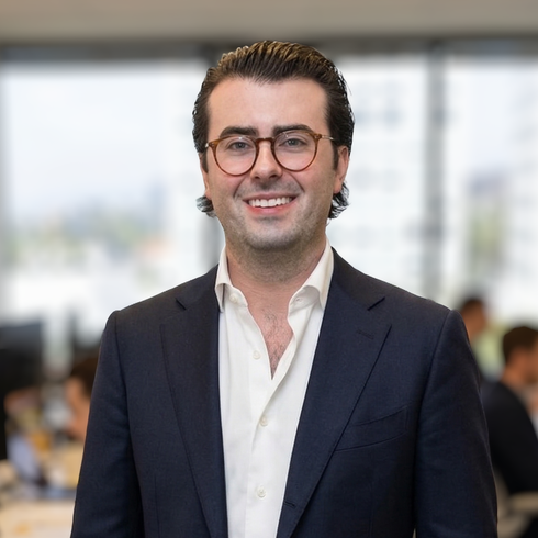 A professional headshot of a person wearing round-frame glasses and a navy blazer with an open-collared white shirt.