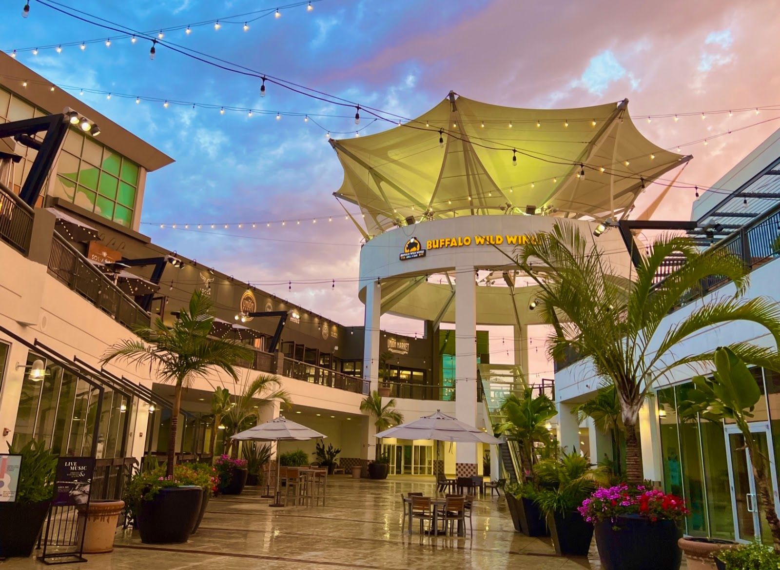 Outdoor plaza at dusk with a large, white canopy, string lights, and potted tropical plants under a colorful sky.