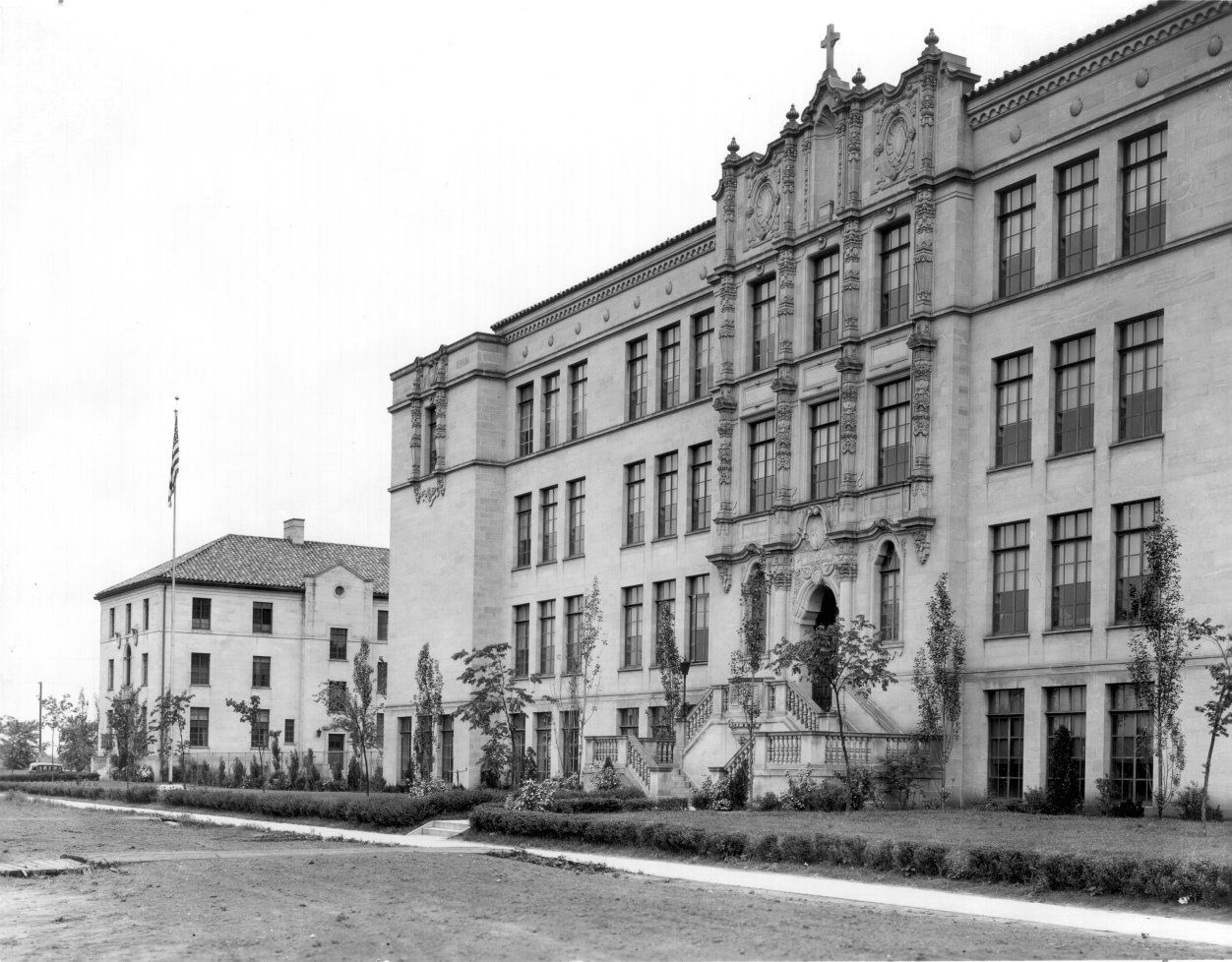 A black-and-white view of a large, multi-story masonry school building with ornate stone detail and a smaller adjacent hall.