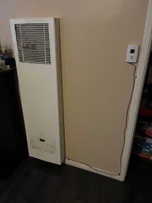 White wall heater beside a beige wall with a small thermostat and exposed cord near the floor.