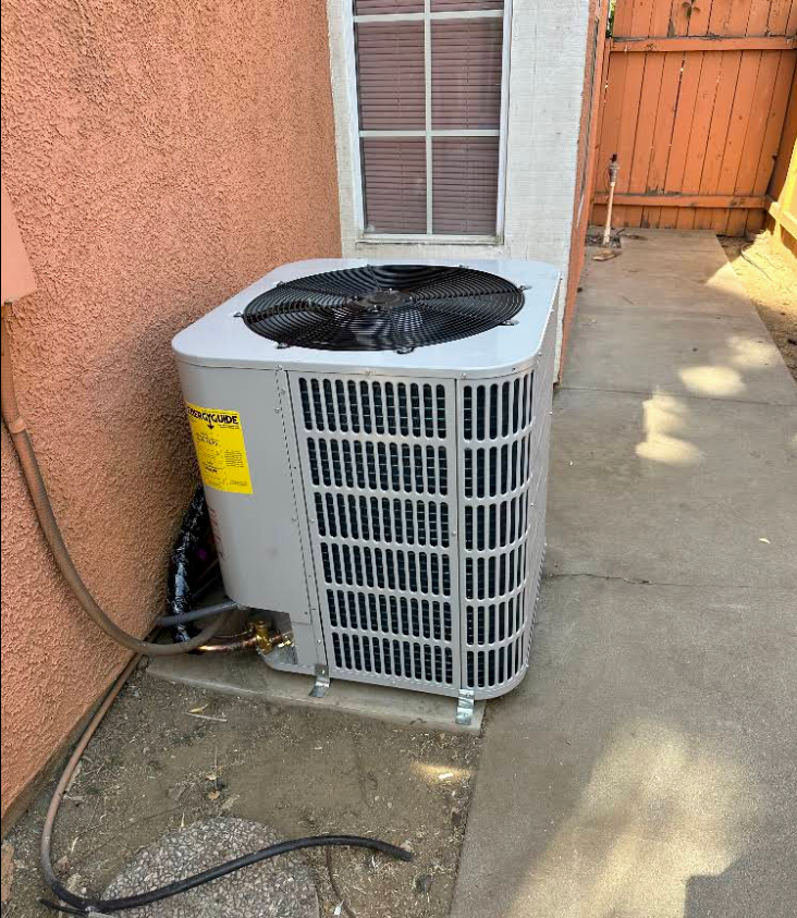 Outdoor air conditioning unit beside a peach stucco wall and window, with exposed hoses on concrete.