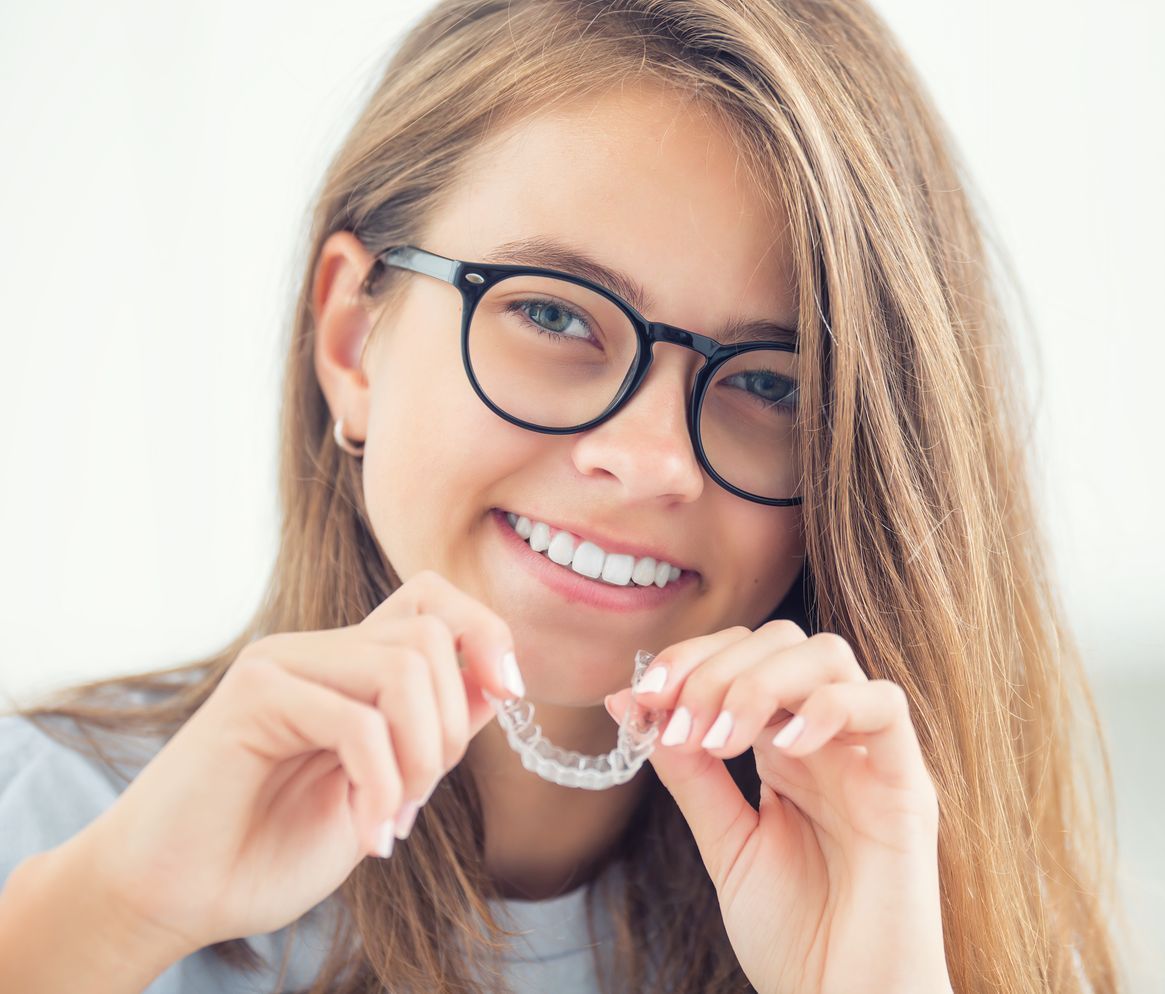 A young woman wearing glasses is smiling while holding a clear retainer.