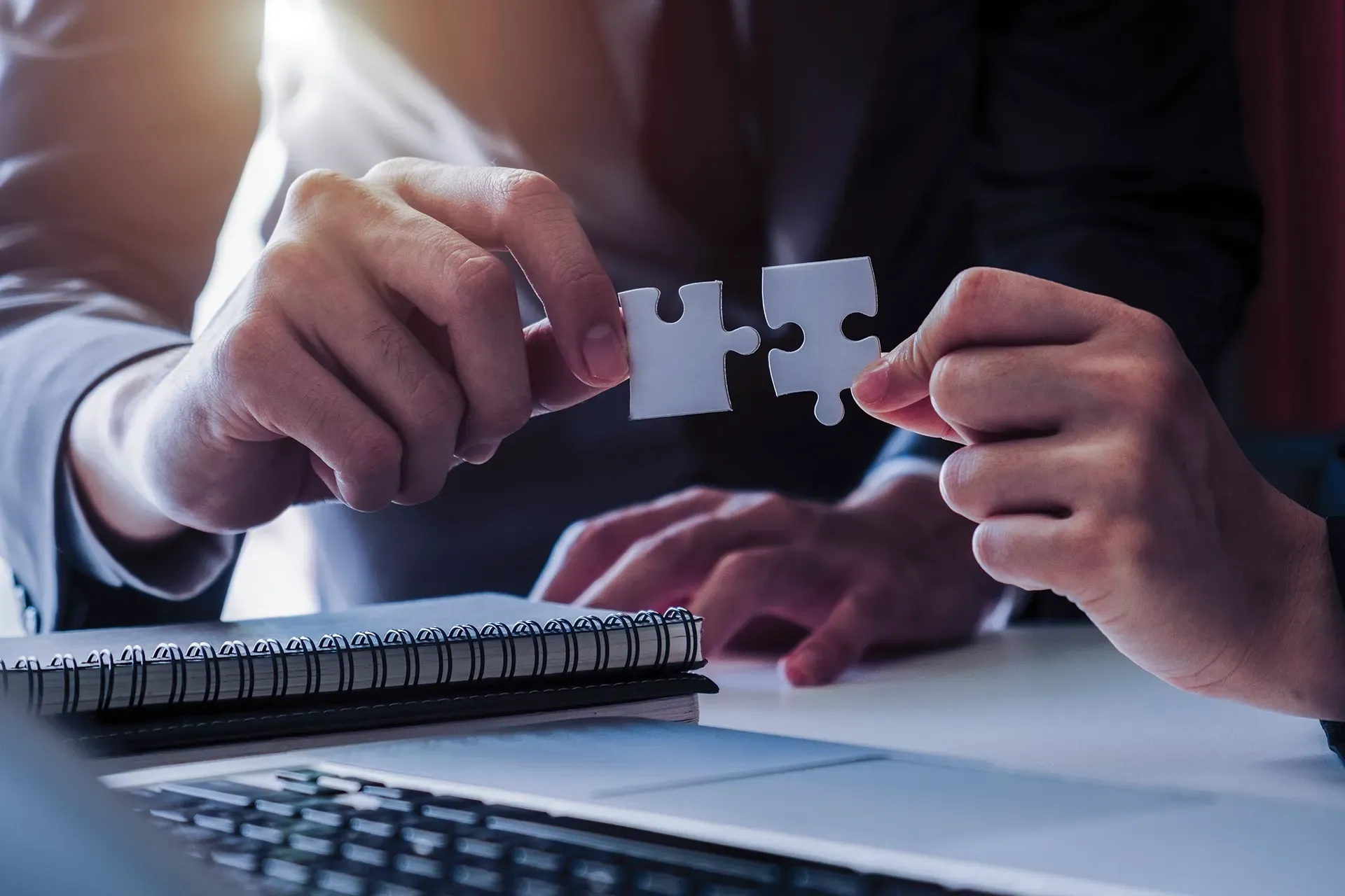 Two people fitting puzzle pieces together over a desk, symbolizing collaboration and problem-solving.