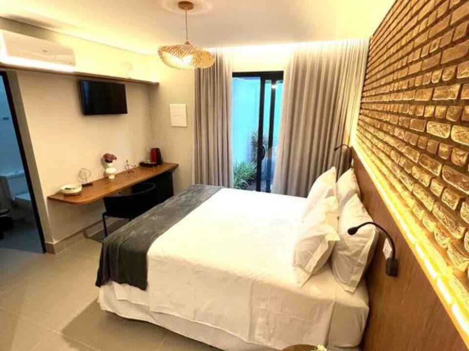 Quarto de hotel moderno com cama, escrivaninha, TV de parede e porta de correr para uma varanda.