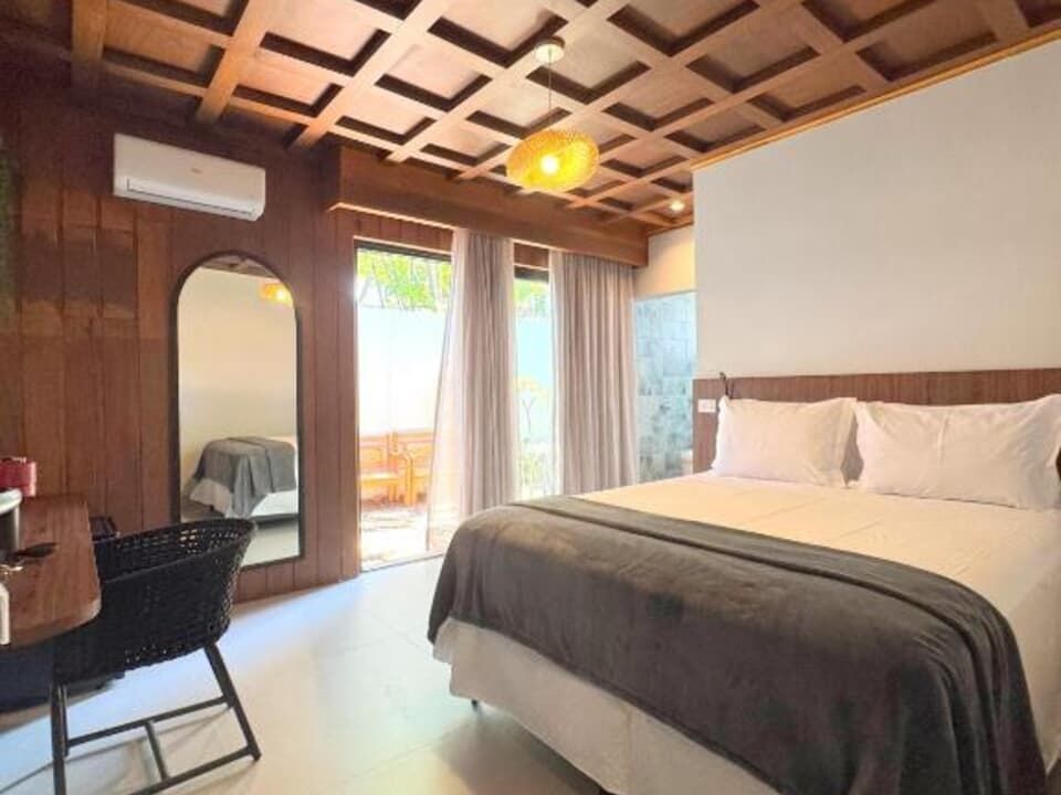 Quarto moderno com cama king-size, teto de madeira, escrivaninha, espelho e portas de sacada com cortinas translúcidas.