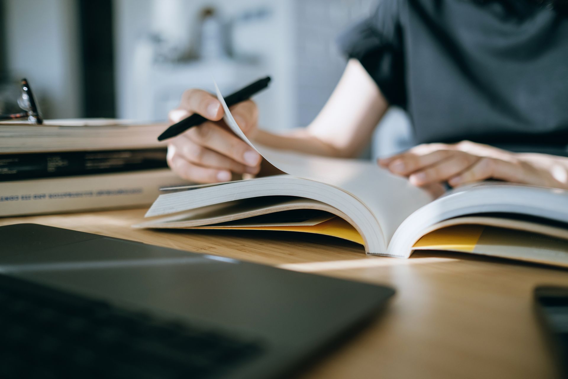Person flipping pages of a book, with a pen, laptop, and stacked books on a wooden table.