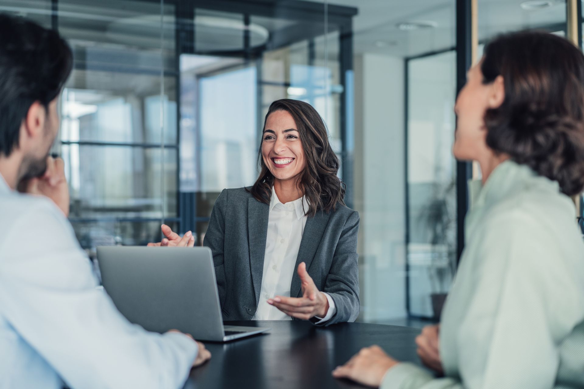 Woman in a suit smiles and gestures, talking to a couple at a desk with a laptop in an office setting.