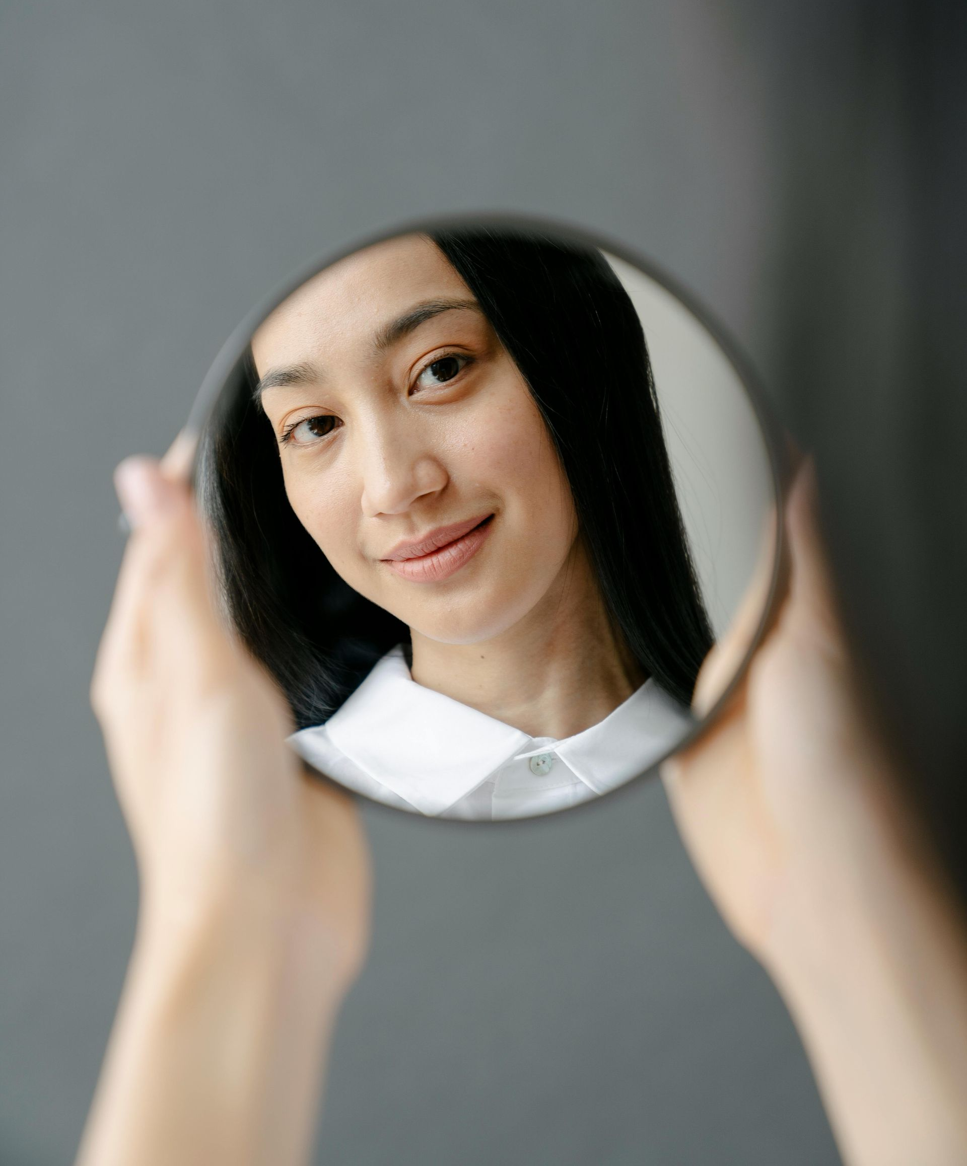Manos sosteniendo un espejo redondo que refleja a una persona con cabello largo y negro y una camisa de cuello blanco.