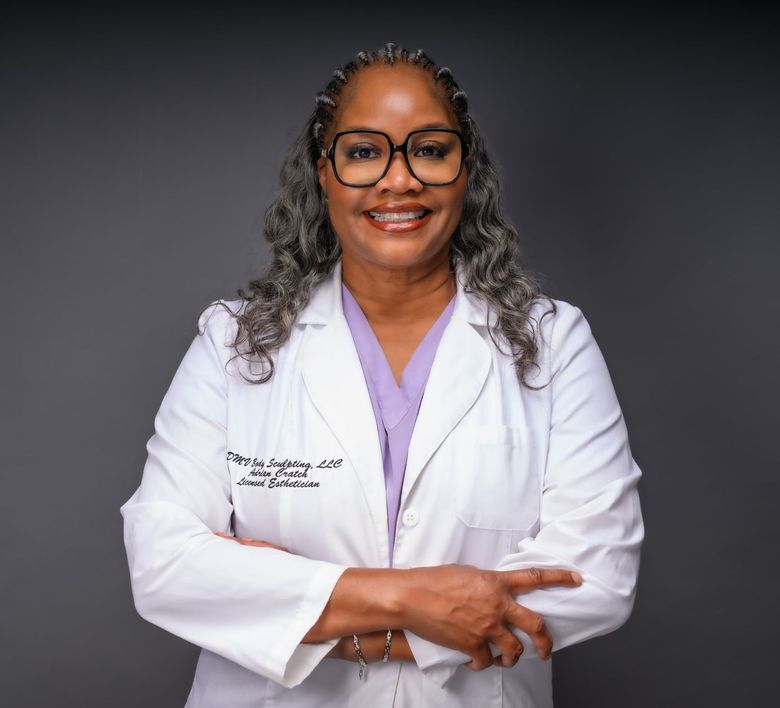 A smiling woman with blonde hair wearing a white lab coat. Gold hoops, neutral background.