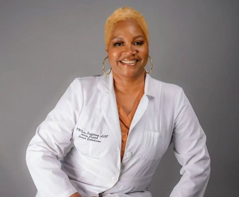 A smiling woman with blonde hair wearing a white lab coat. Gold hoops, neutral background.