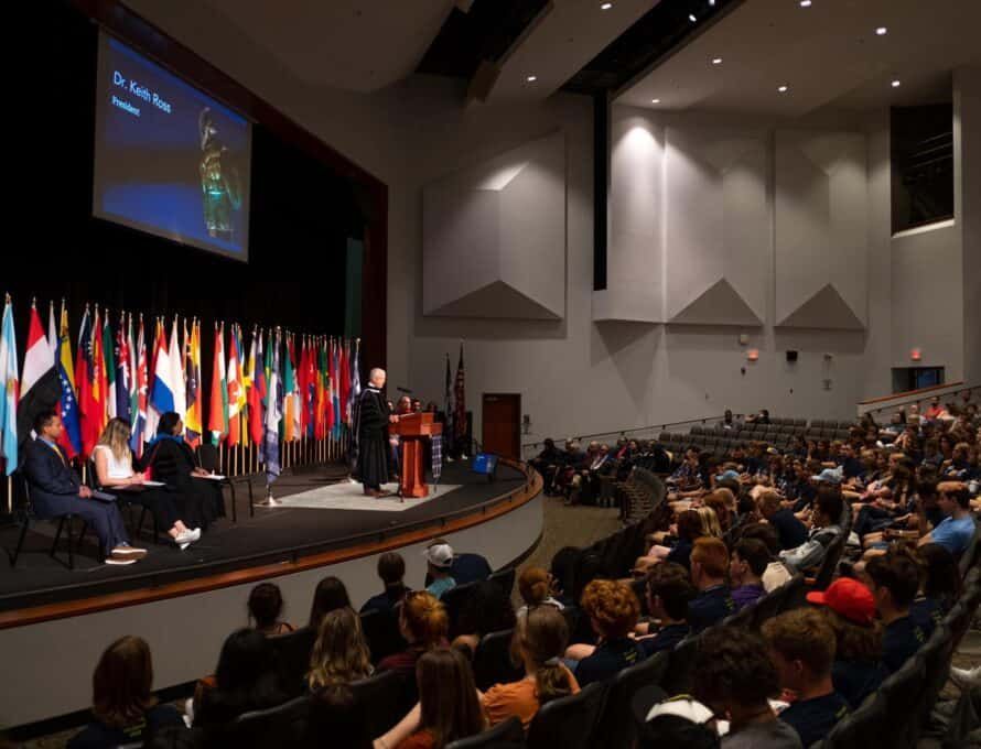 Auditorium with a presentation; flags line the stage. A speaker addresses a seated audience.