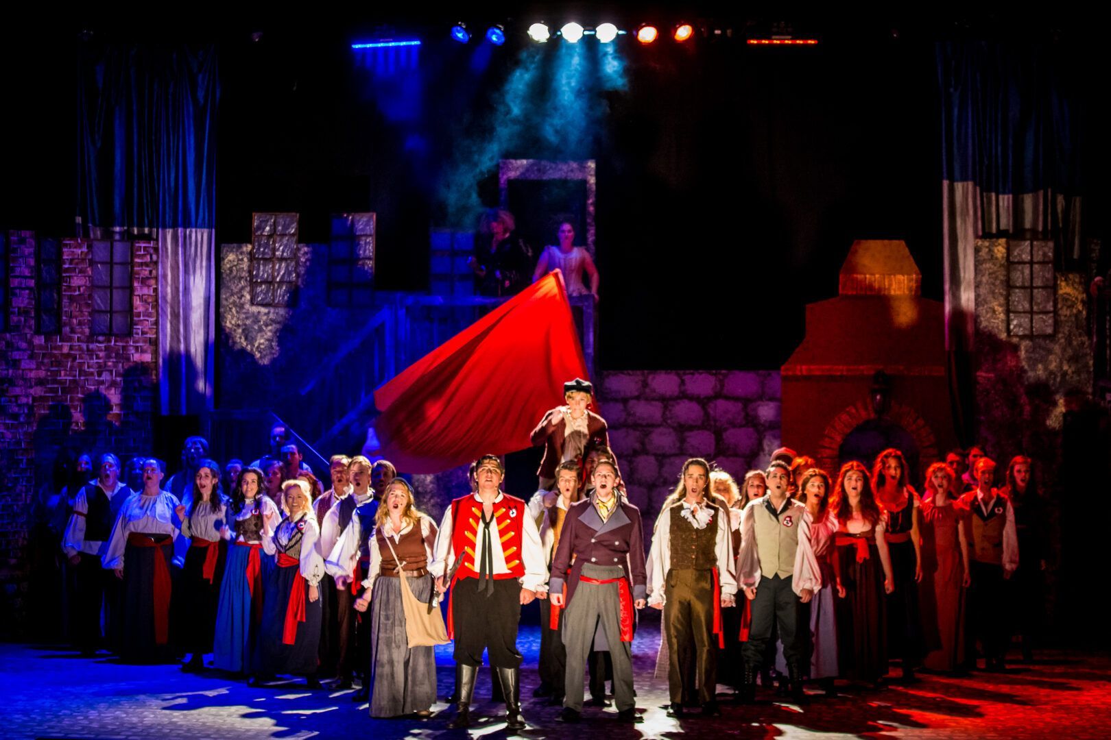 Auditorium AV solutions. Stage production of a musical with actors in costume, red, blue stage lighting, and a large cape.