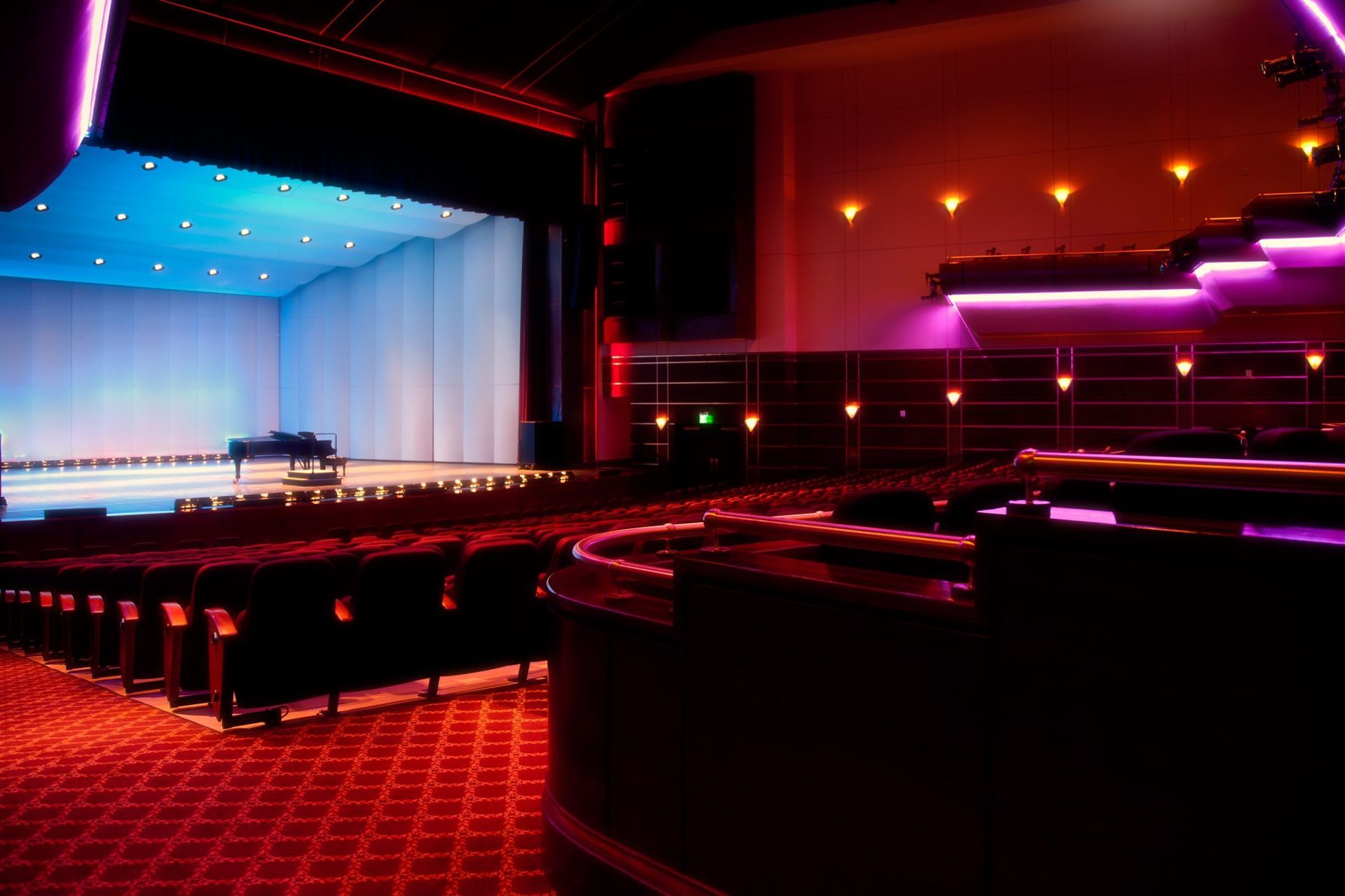Auditorium AV solutions. Interior of a theater with red carpet and seating, stage with piano and blue lighting.