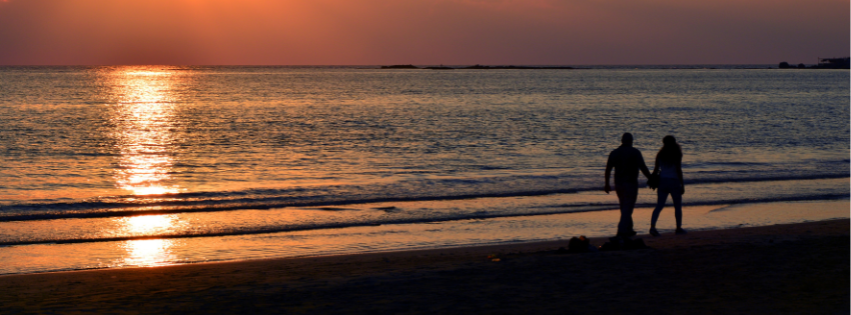 A man and a woman are walking on the beach at sunset holding hands.