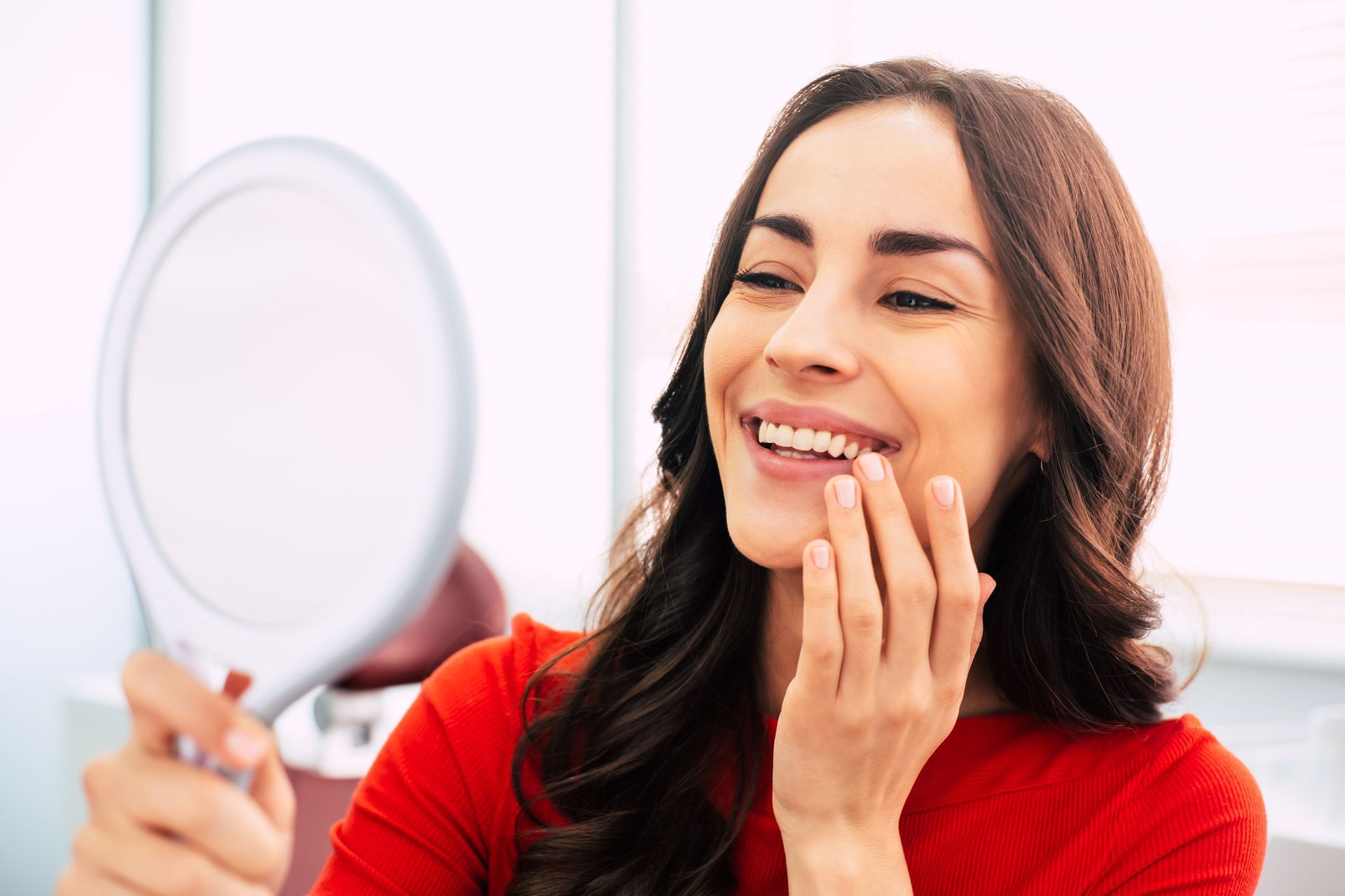 Woman in red shirt smiling, examining teeth in a hand mirror.