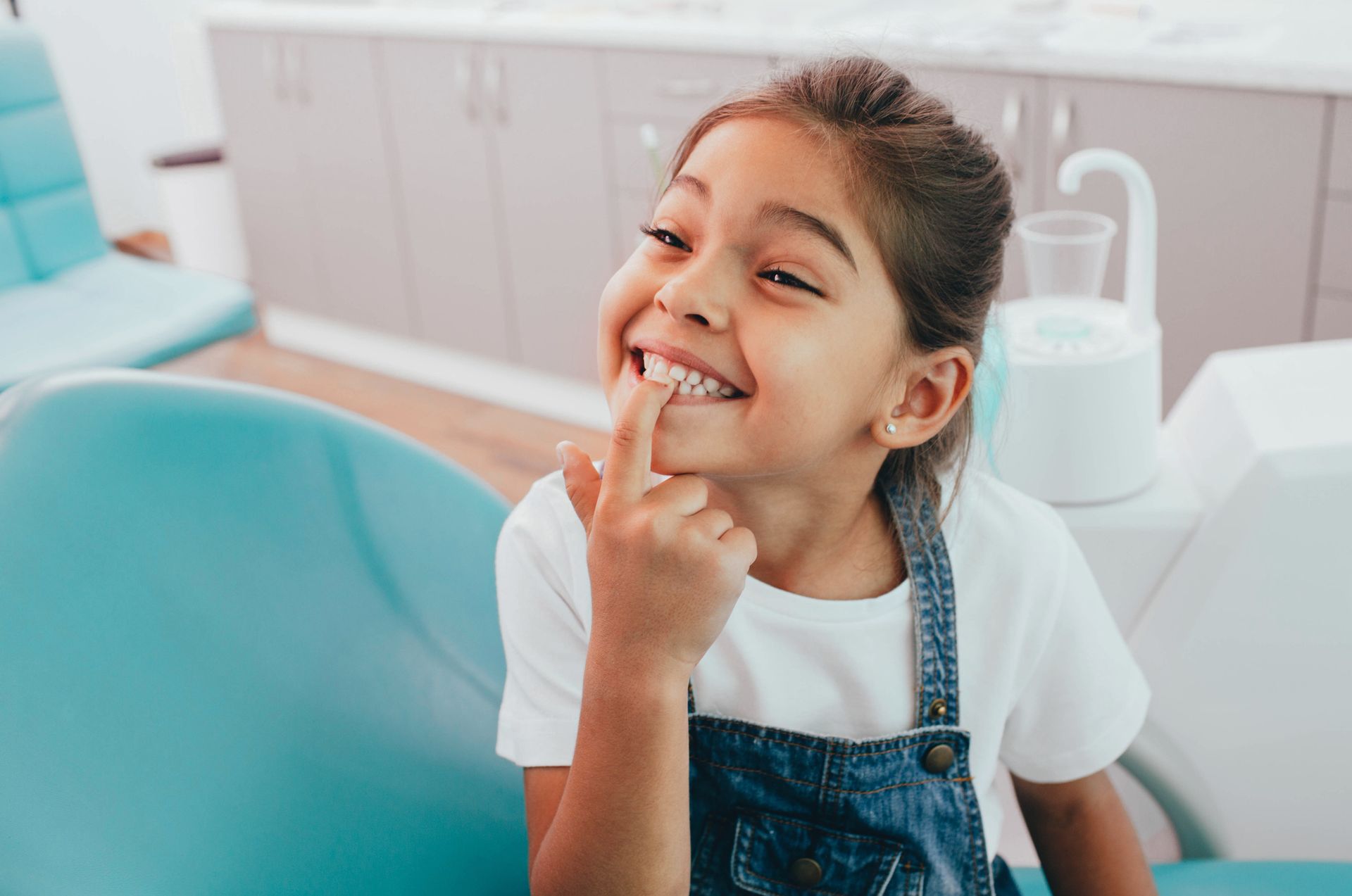 Smiling girl at a dentist's office points to her teeth. Blue chair, white shirt, denim overalls.