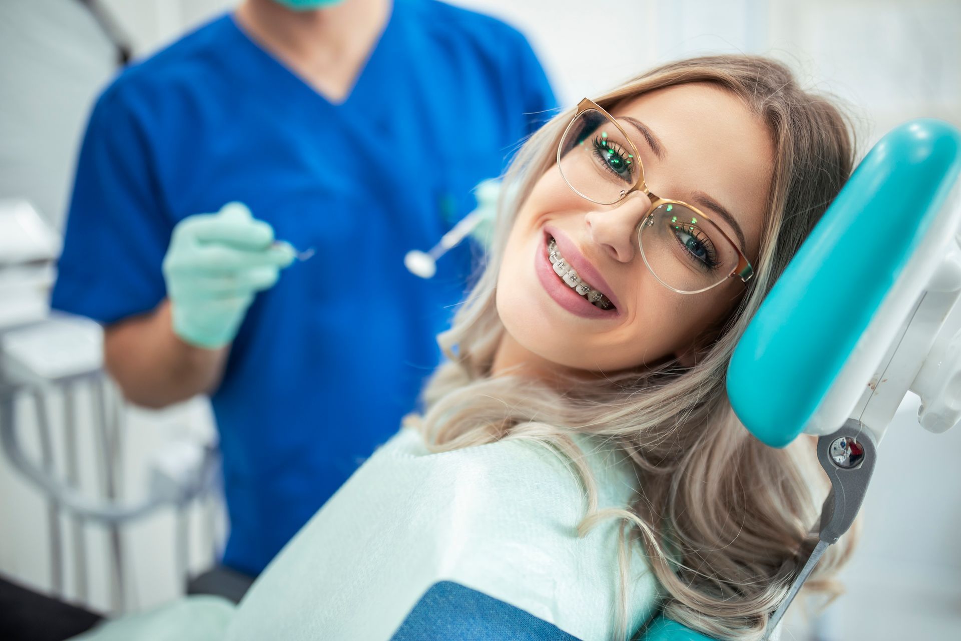 Woman with braces smiles in a dental chair, a dentist in blue scrubs behind her.