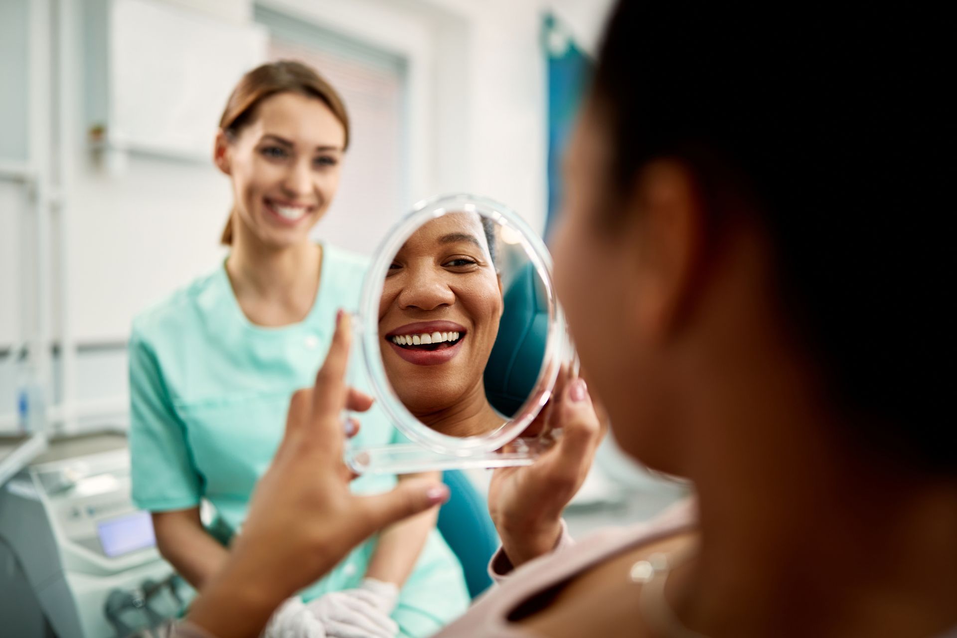 Woman in dental chair smiles in mirror, dentist in green scrubs looks on.