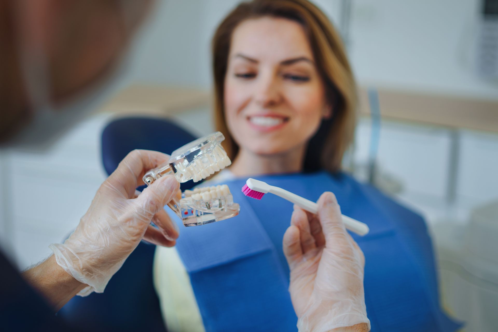 Dentist showing a model of teeth to a smiling woman in a dental chair, demonstrating brushing technique.