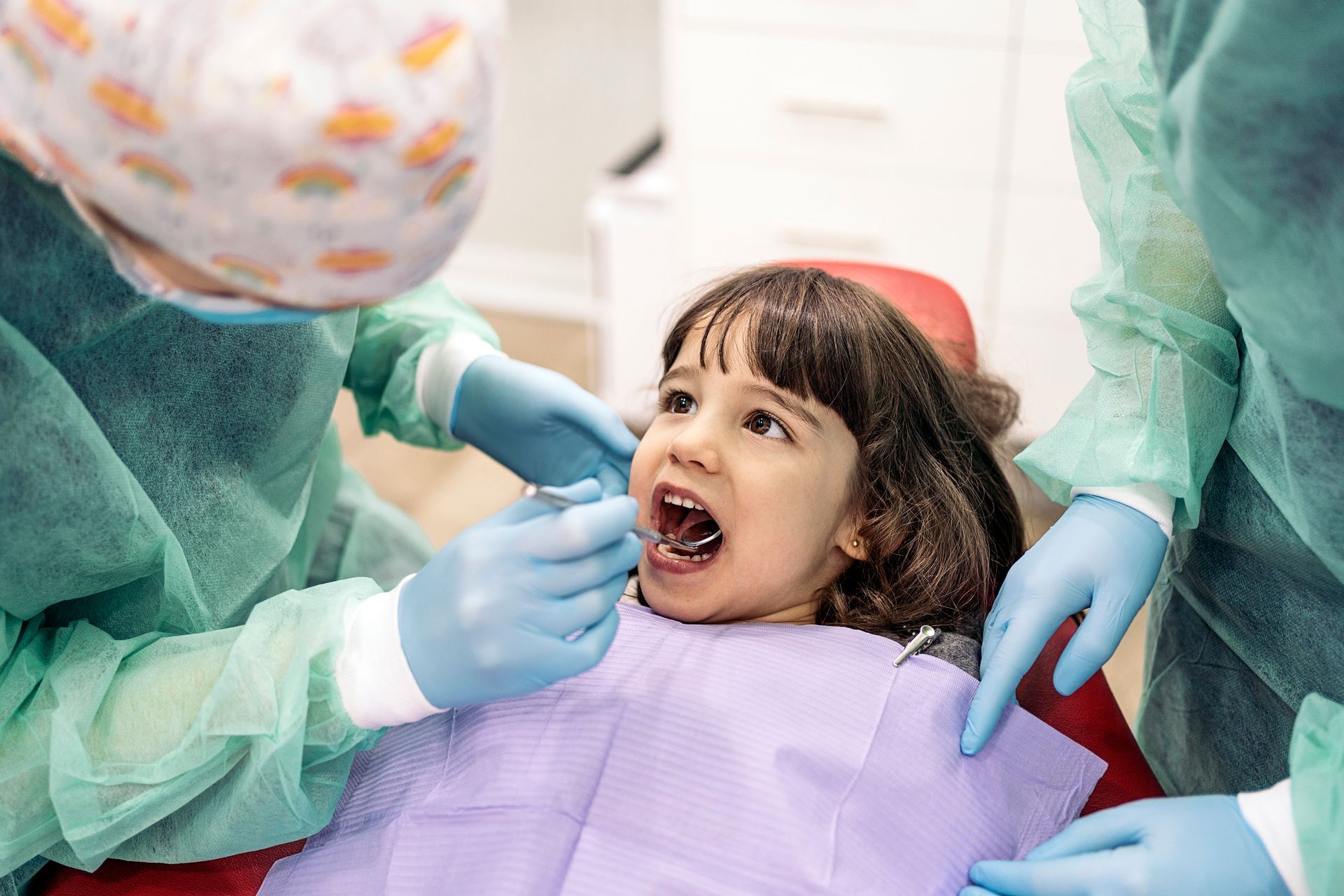 Child at the dentist, mouth open, being examined. Two dentists in scrubs and gloves.