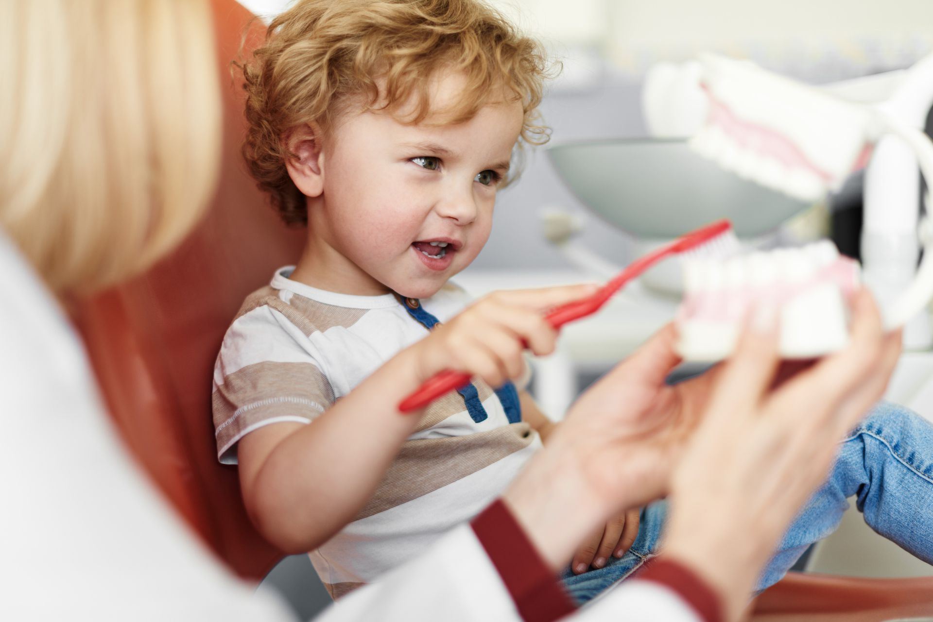 Young child in dentist chair practicing brushing teeth with a model under supervision.