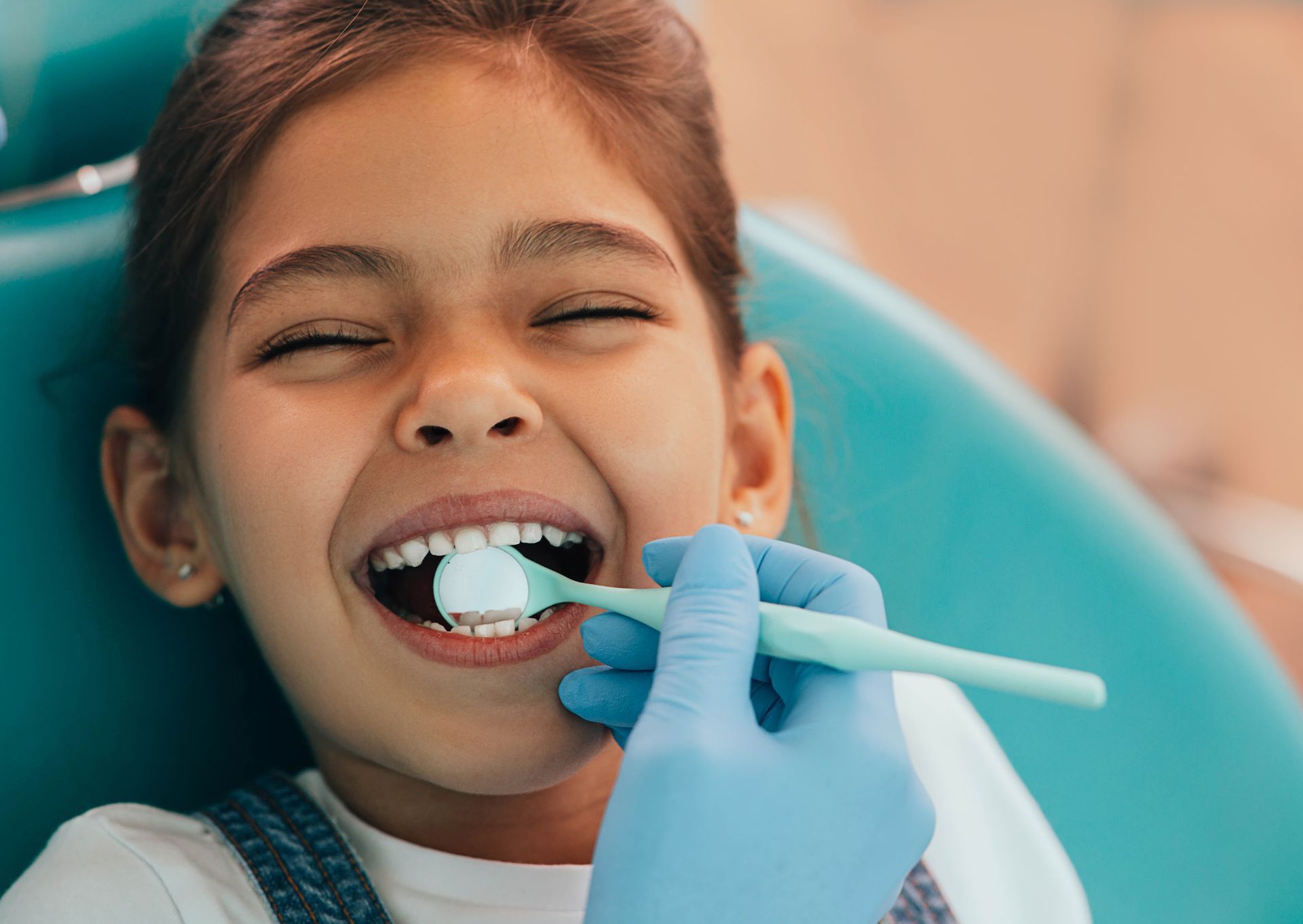 Girl at dentist, mouth open, dentist examining teeth with a mirror in a clinic, blue chair, gloved hand.