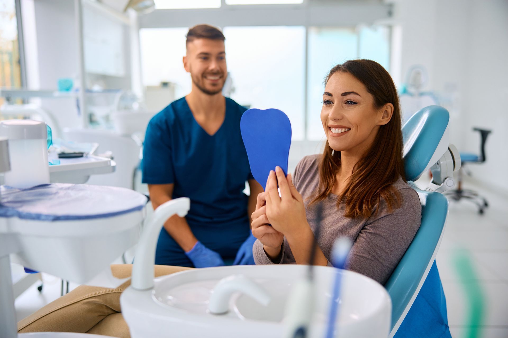 Woman in dentist chair examines teeth in mirror, smiling. Dentist in blue scrubs smiles.