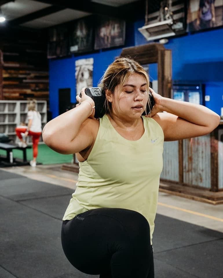 A woman is doing squats with a dumbbell on her head in a gym.