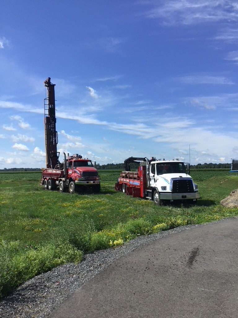 Deux camions de forage sur un champ herbeux sous un ciel bleu. Un rouge, un blanc.