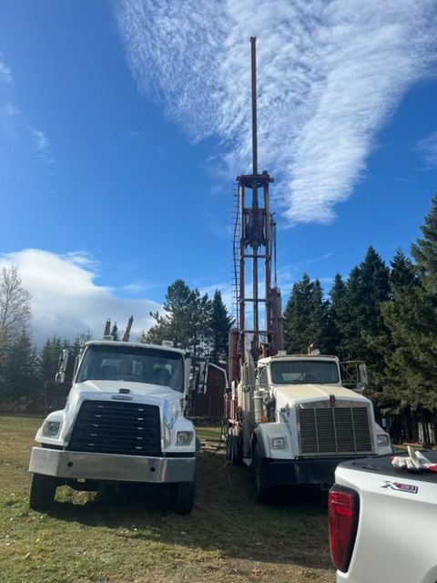 Deux camions blancs et une foreuse sur un terrain herbeux, sous un ciel bleu nuageux.