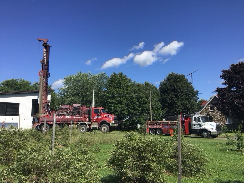 Une foreuse et deux camions sur un terrain herbeux, prêts à forer près d'un bâtiment. Ciel d'un bleu éclatant.