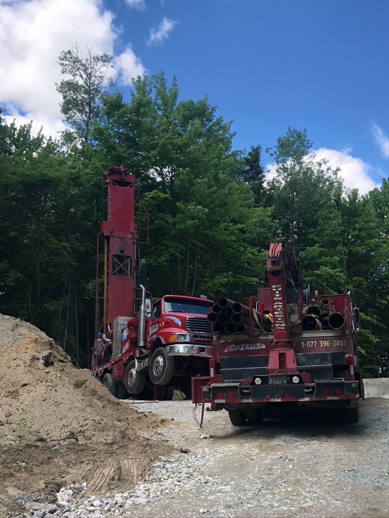 Une foreuse rouge sur un chemin de gravier, à côté d'une remorque chargée de tuyaux, devant une forêt sous un ciel bleu.
