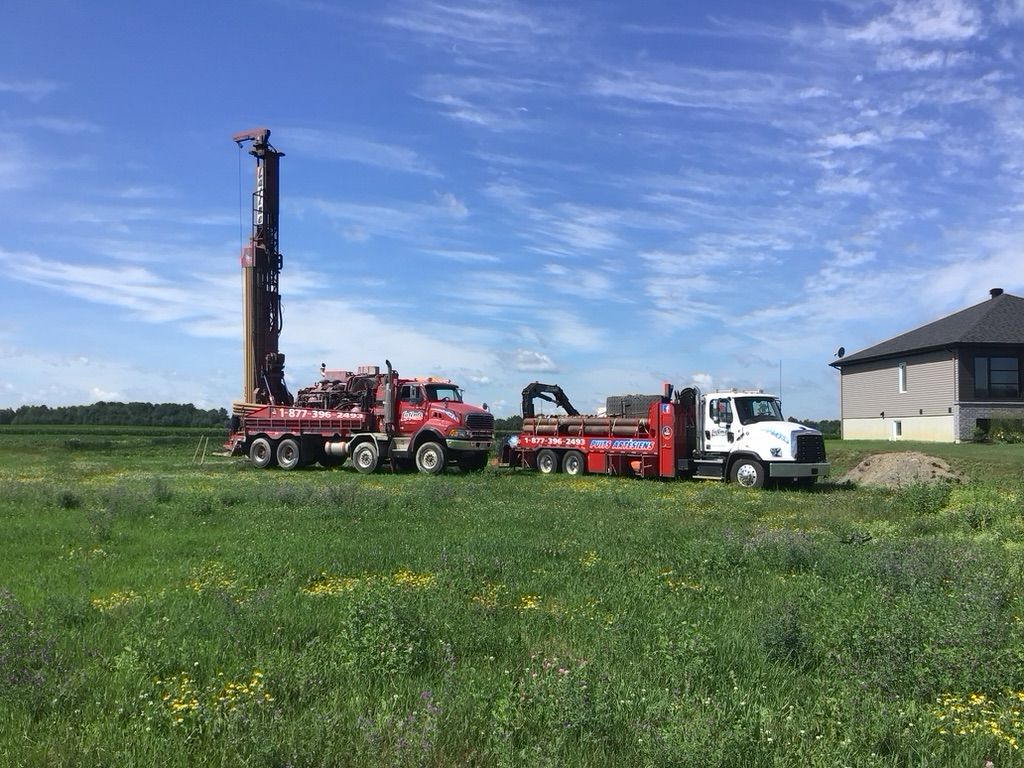 Un camion de forage rouge et un véhicule d'assistance forent un puits près d'une maison sous un ciel bleu, dans un champ herbeux.