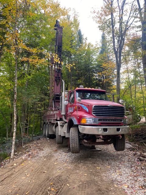 Camion de forage rouge sur un chemin de terre dans une zone boisée.