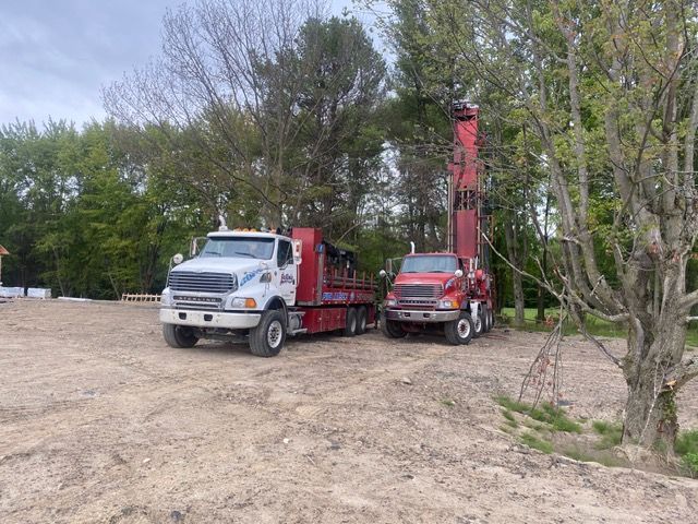 Deux camions : un blanc et une foreuse rouge, garés sur un terrain vague avec des arbres en arrière-plan.