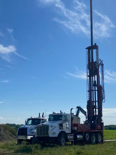 Deux camions dans un champ, l'un d'eux transportant du matériel de forage en hauteur. Ciel bleu avec des nuages en arrière-plan.