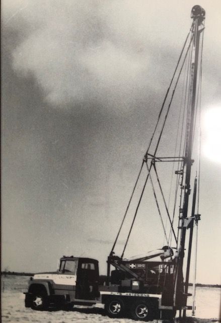 Photo en noir et blanc d'une foreuse de puits montée sur un camion dans un champ, sous un ciel nuageux.